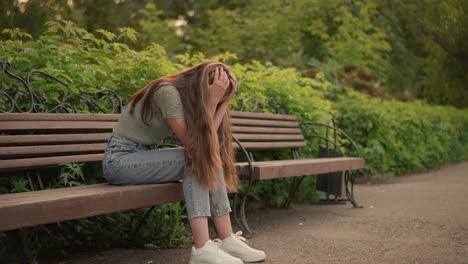 side view of woman sitting on wooden bench, hands gently covering her face in reflective, somber posture, her long hair cascading down her shoulder, surrounded by lush greenery