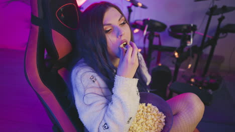 top down view of young lady with painted eye makeup seated on gaming chair eating popcorn while focused on something out of frame with electronic drum set softly blurred in background