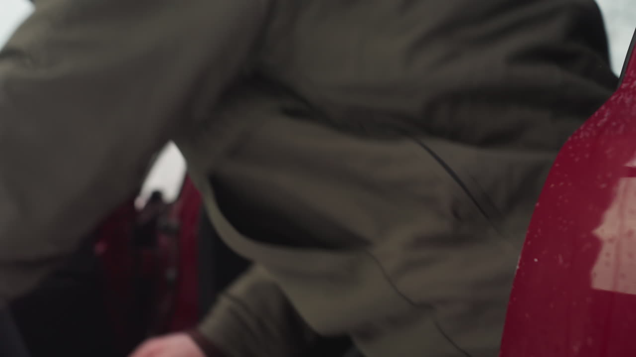 Man wearing sunglasses steps out of red car during light snowfall with visible snow-covered background, vehicle interior in view, and water droplets on car door