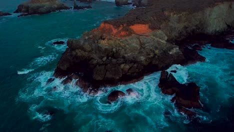 Slow-orbit-around-rocky-ocean-point-deluged-by-crashing-waves-at-Sand-Dollar-Beach-in-Big-Sur-California-at-dusk