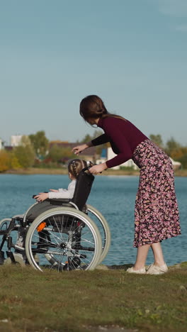 woman and child with spinal cord injury rest on riverbank. couple discusses houses and plants on opposite bank pointing with fingers backside view