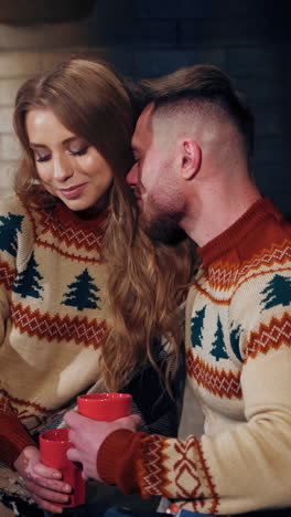 beautiful loving couple resting near fireplace. young woman and a man with cups of drink at christmas. guy hugging his girlfriend enjoying romantic weekend. vertical video