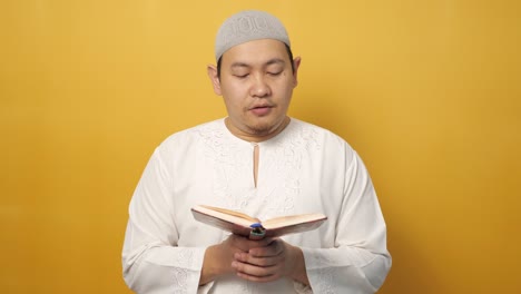 젊은 아시아 무슬림 남성의 초상화, 노란색 배경에 대한 거룩한 꾸란을 읽는, 이슬람 주제, 라마단 카림 또는 이드 무바라크