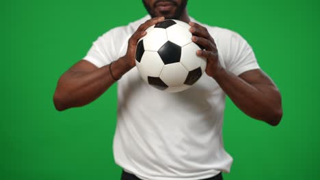 unrecognizable serious african american footballer holding ball in hands standing at chromakey background. front view concentrated confident sportsman on green screen playing soccer.
