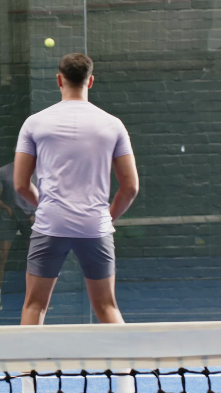 Vertical video: Tossing ball, man in purple tee swinging paddle on blue glass court, practicing