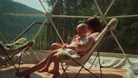 mujer sostiene a un niño encantador en los brazos sentado en un balcón de vidrio. madre e hijo se relajan en un sillón a la luz del sol mirando la vista desde una amplia ventana. concepto de naturaleza increíble