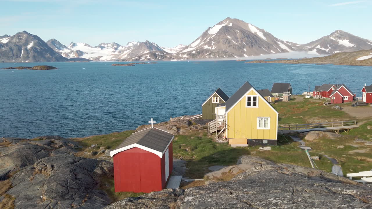 Fredelig og fargerik kystlandsby på Grønland med snødekte fjell i det fjerne og små hus i forgrunnen med en liten rød kirke.