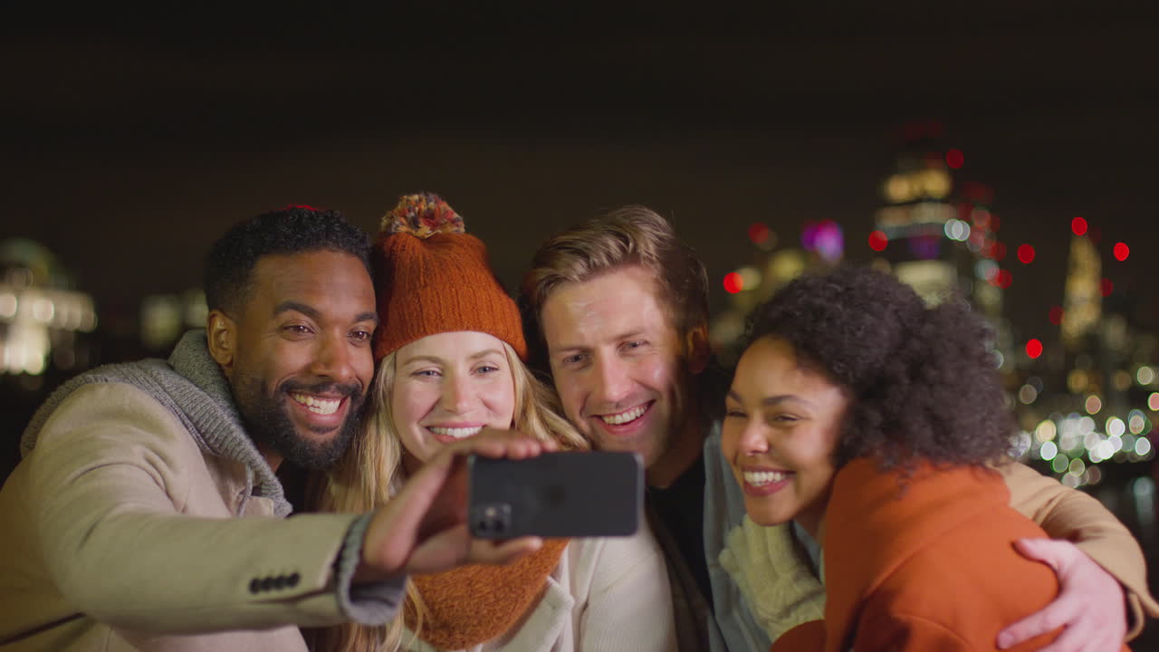 grupo de amigos con abrigos de invierno y bufandas posando para una selfie en el teléfono móvil con las luces de la ciudad en el fondo - filmado en cámara lenta