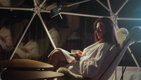 mujer equilibrada descansando iluminada por la luz de la linterna disfruta del café de la noche después del día de trabajo. mujer joven sentada en el sillón con calma mira lejos en una atmósfera romántica