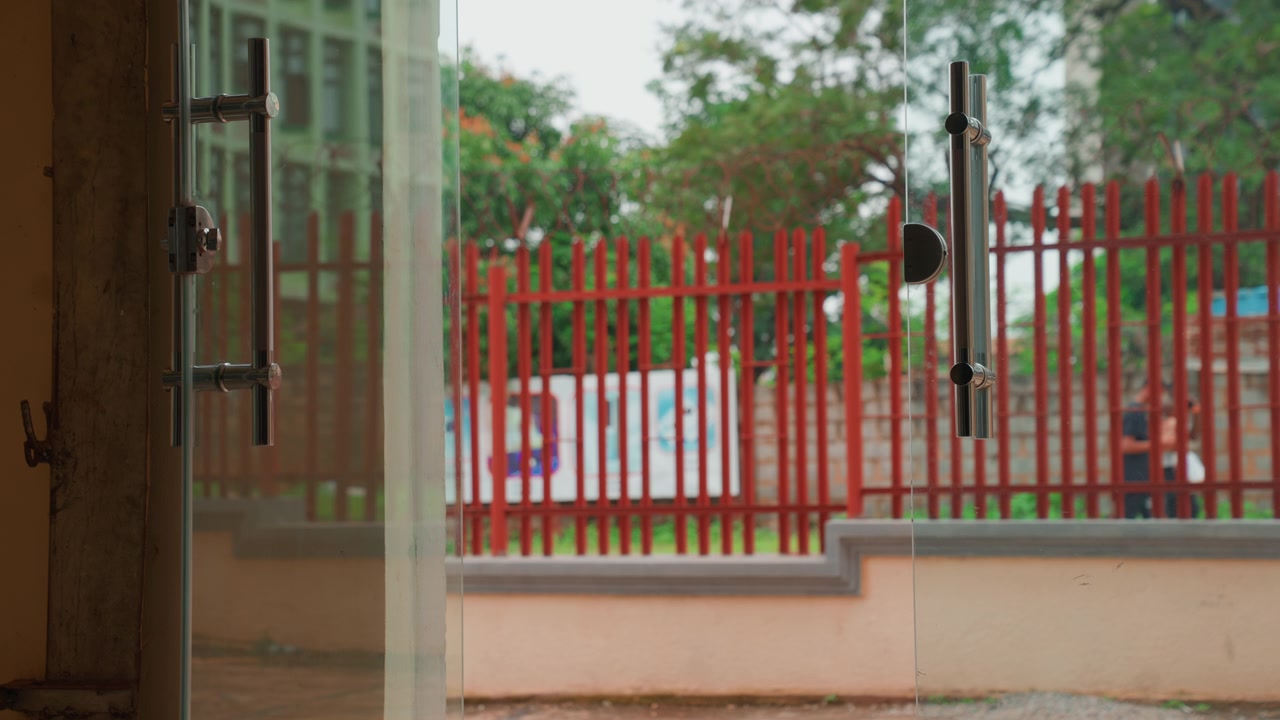 Open glass door framing red fence and quiet street, interior threshold with metal handle and latch in foreground, exterior yard and trees beyond fence, muted color palette and soft light, calm waiting