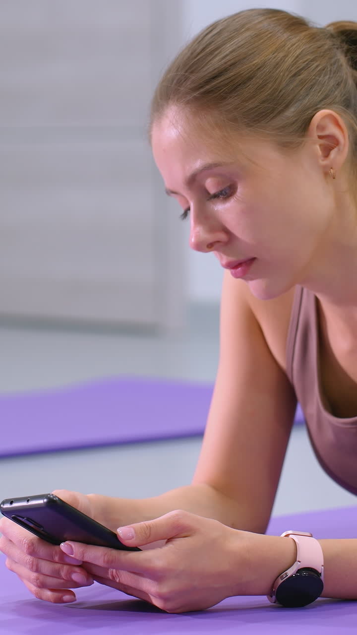 joven enfocada yace en una alfombra de yoga púrpura, opera un teléfono inteligente mientras usa un reloj inteligente, vestida con ropa atlética, parece relajada en un estudio de fitness, el fondo presenta una decoración minimalista