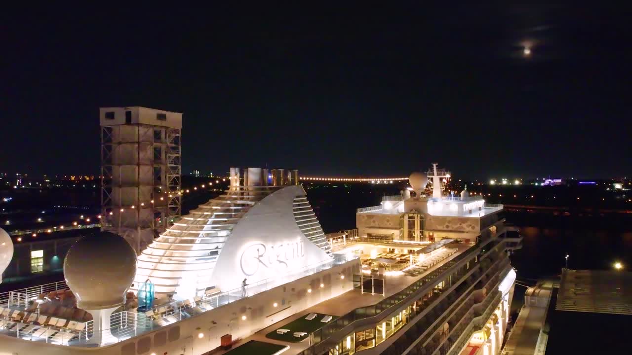 The Regent Seven Seas Grandeur cruise ship docked in port, beautifully lit against the night skyline under full moon, sparkling lights against dark sky