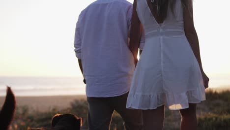 Toma-Manual-En-Cámara-Lenta-De-Una-Pareja-Enamorada-De-La-Mujer-Vestida-Con-Un-Vestido-Blanco,-Su-Hombre-Vestido-Con-Camisa-Blanca-Con-Su-Perro-Durante-Un-Paseo-Romántico-En-La-Playa-En-Una-Hermosa-Puesta-De-Sol