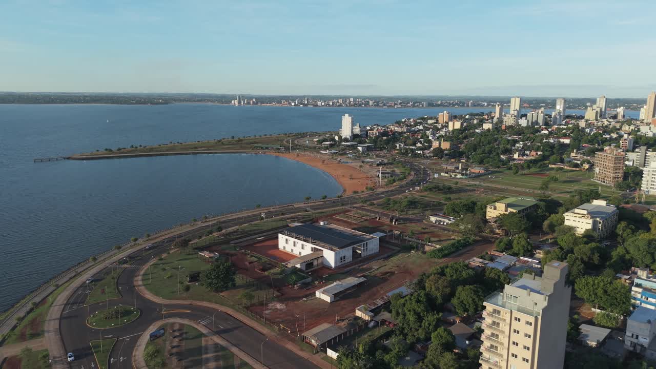 Drone view of Posadas, Misiones, Argentina, features Bahía El Brete, waterfront boulevard, urban buildings, green areas, and the Paraná River, with visible city infrastructure, real time.