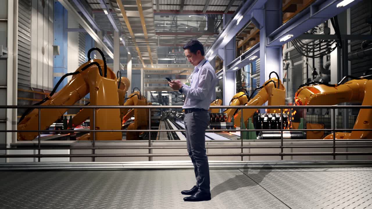 Full Body Side View Of An Asian Male Professional Worker Standing With His phone With Industrial Robotic Arms Working, Working Continuously With His phone