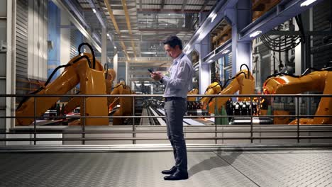 full body side view of an asian male professional worker standing with his phone with industrial robotic arms working, working continuously with his phone