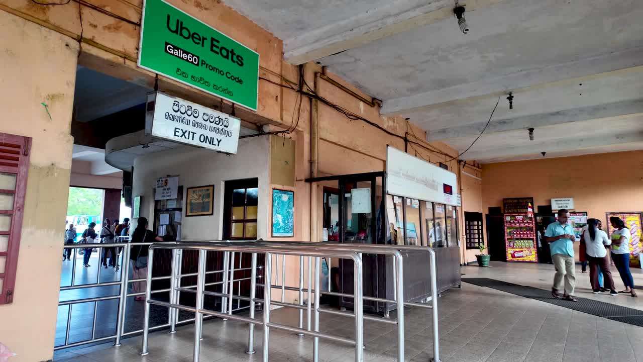 Kandy railway station's exit area featuring a ticket booth, exit sign, and various signage. The scene captures the transport hub's lively atmosphere in Sri Lanka.