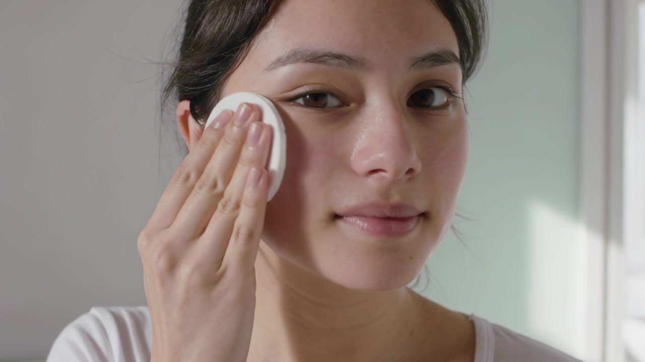 Donna che preme un dischetto di cotone sotto l'occhio sinistro, pulendosi la guancia a casa mentre fa la sua routine di skincare, indossando un top bianco.