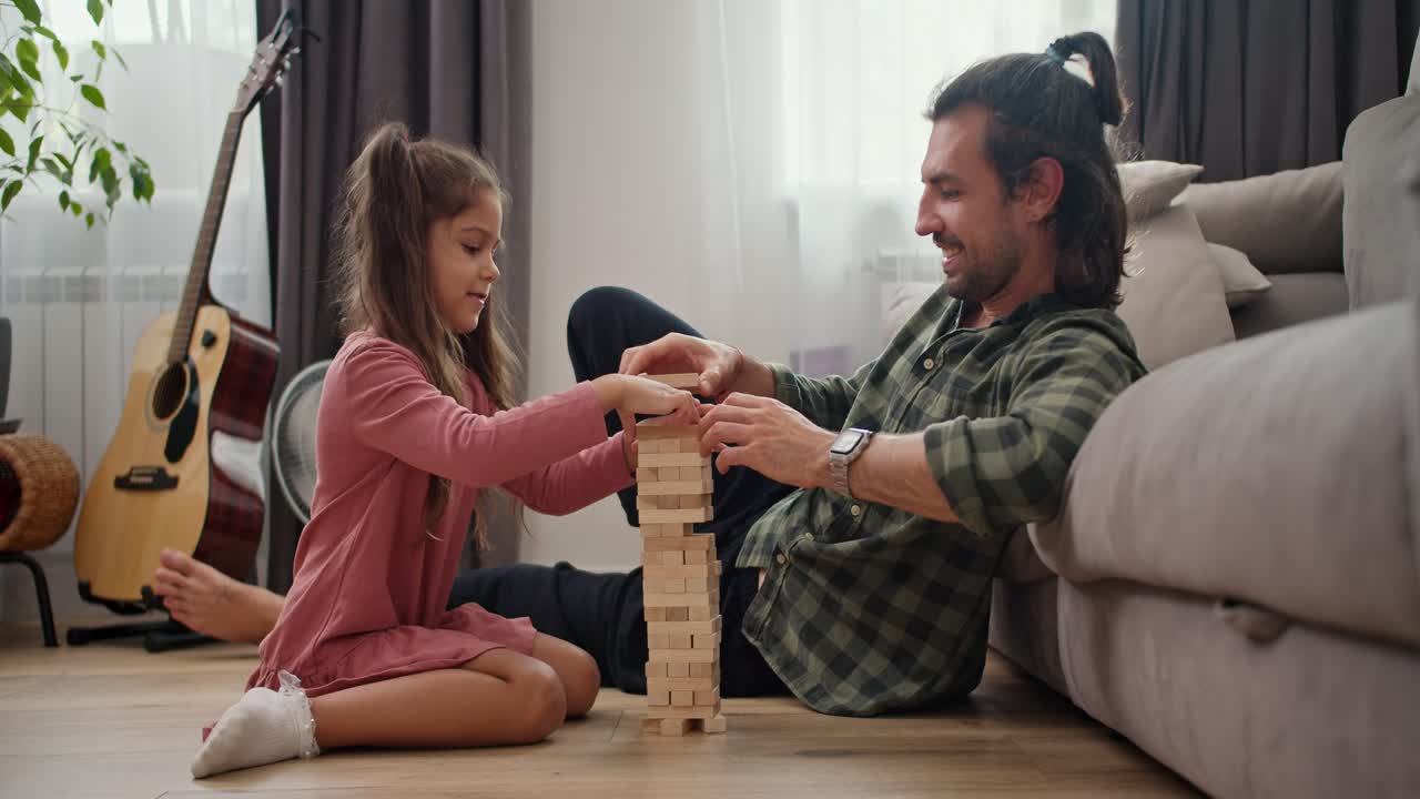 un padre solitario, un hombre moreno con una camisa a cuadros verde juega con su hija pequeña, una chica morena con un vestido rosado, en el juego de mesa jenga, apoyado en un sofá gris en el suelo en un apartamento moderno