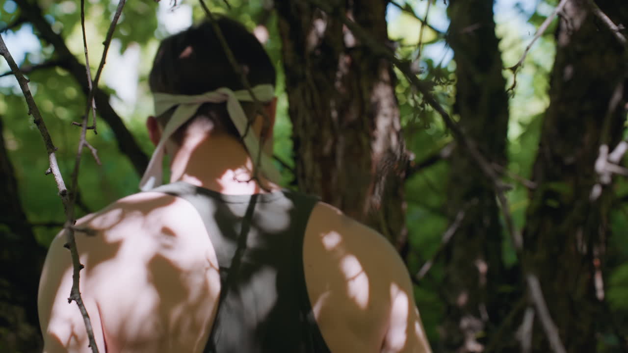 back view of shirtless young man in headband delivering powerful punch and elbow strike to gnarled tree bark, shoulders flexing with effort amid dense tangled branches and mottled sunlight