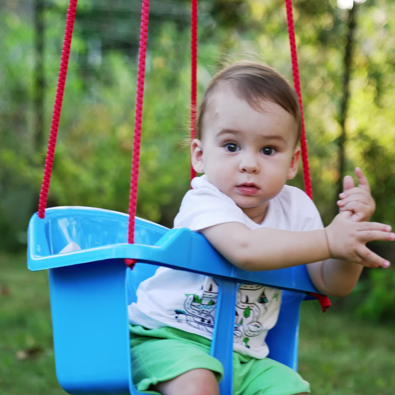 Caucasian baby boy swaying in the blue swing. Lovely child enjoying summertime in the nature. Blurred background