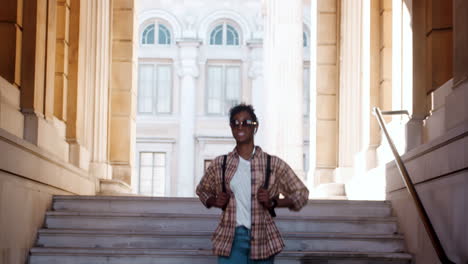 Front-view-of-young-black-woman-wearing-sunglasses,-blue-jeans-and-a-plaid-shirt-walking-down-steps-outside-a-historical-building-looking-around-and-listening-to-music-with-earphones,-low-angle