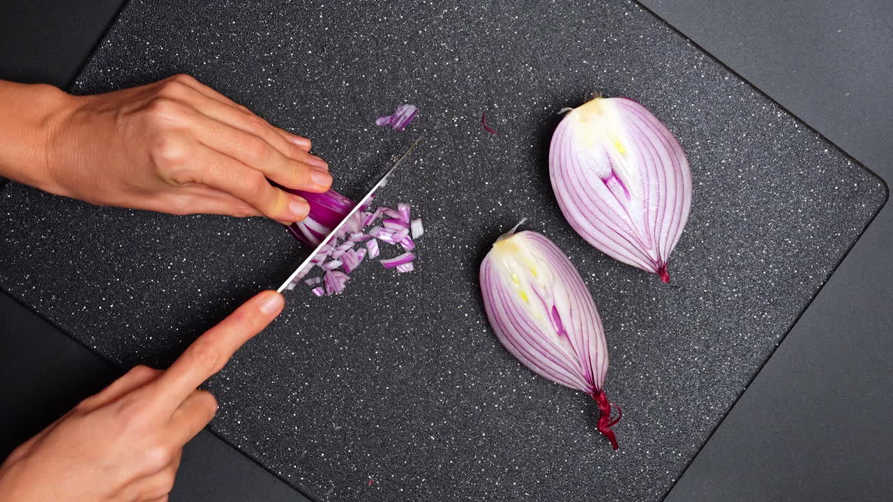 Chopping onions on a black cutting board in a kitchen setting. Hands are cutting red onions on a cutting board in a kitchen