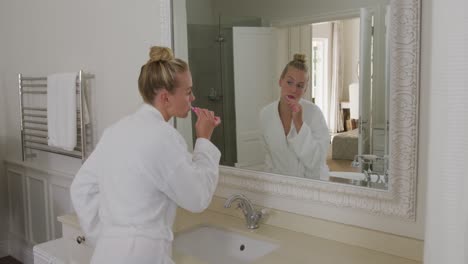 Caucasian-woman-in-bathrobe-brushing-her-teeth-while-looking-in-the-mirror-in-the-bathroom