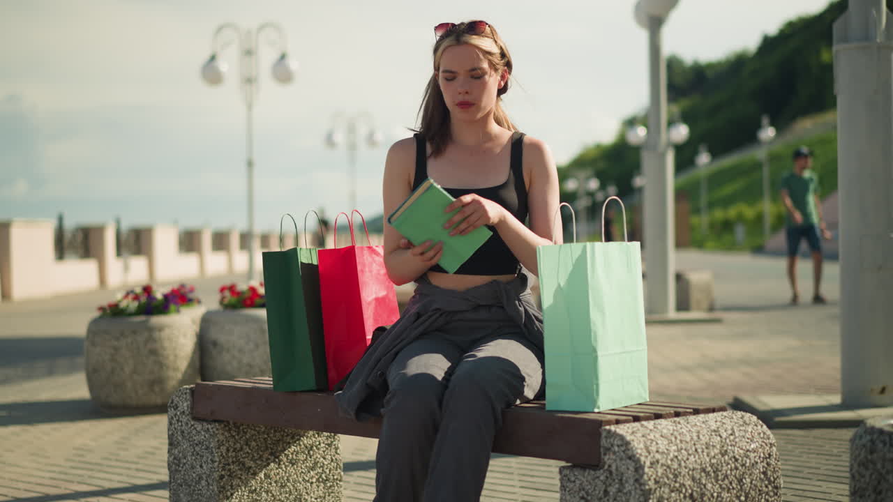 una mujer sentada en un banco al aire libre con bolsas de compras a ambos lados recupera un libro de la bolsa verde menta, lo abre y hojea sus páginas, con una vista borrosa de personas caminando en el fondo