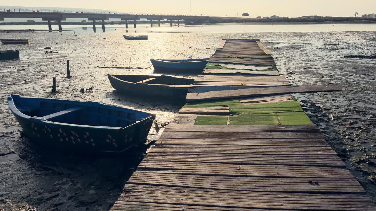 Cinematic scene of walking over old and decayed wooden promenade with dried up and muddied ground surface, old fishing boat and tires at river empty bottom