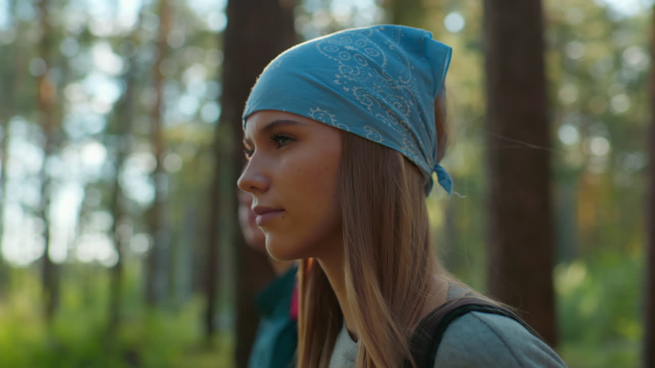 Friends walking through lush green forest with one wearing blue scarf, illuminated by sunlight reflecting off her hair, the lady on green top is softly blurs in the background