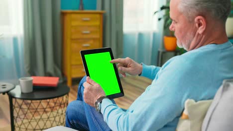 un hombre mayor en casa usa una tableta de pantalla verde. está sentado en un sofá en su acogedora sala de estar. un hombre mayor y guapo hace clic una vez en la pantalla y navega por el contenido