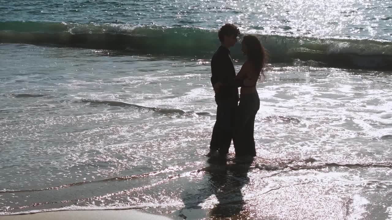 Young Couple Embracing While Standing On Sandy Beach With Waves Splashing. Muxia, Spain. orbiting shot