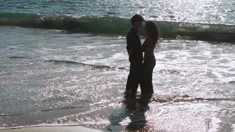 young couple embracing while standing on sandy beach with waves splashing. muxia, spain. orbiting shot