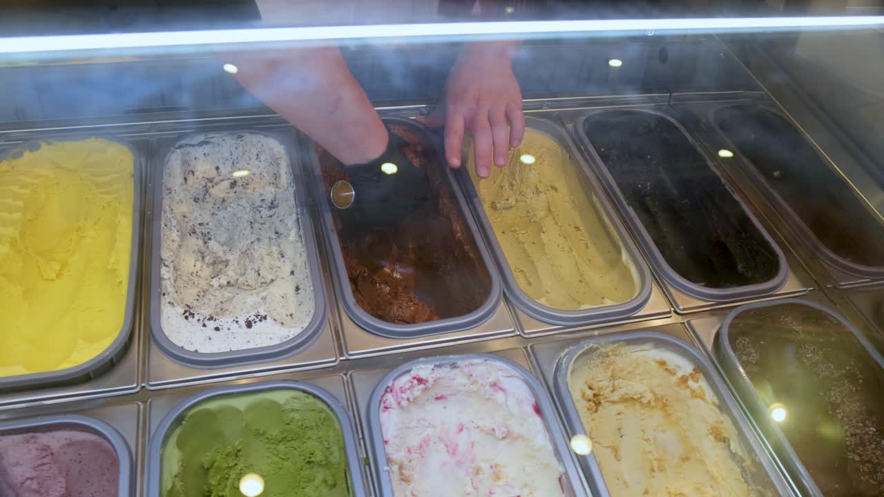 A gloved hand selects and scoops ice cream from a brightly lit display freezer, viewed from above, with multiple colorful flavors visible in metal trays