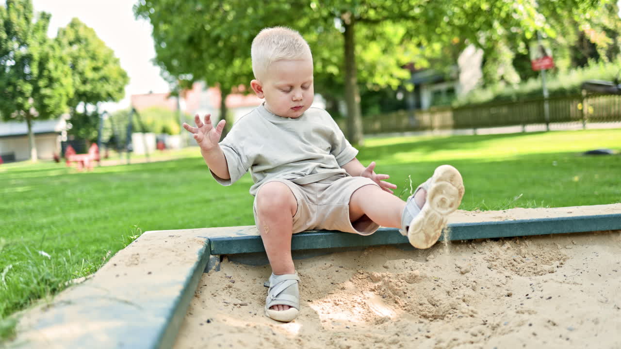 Un bambino allegro in maglietta e pantaloncini scava la sabbia mentre è seduto in una sabbiera. Un bambino piccolo in una sabbiera in un parco giochi. Un bambino per strada scava la sabbia con una paletta di plastica. Giocare con la sabbia per strada.