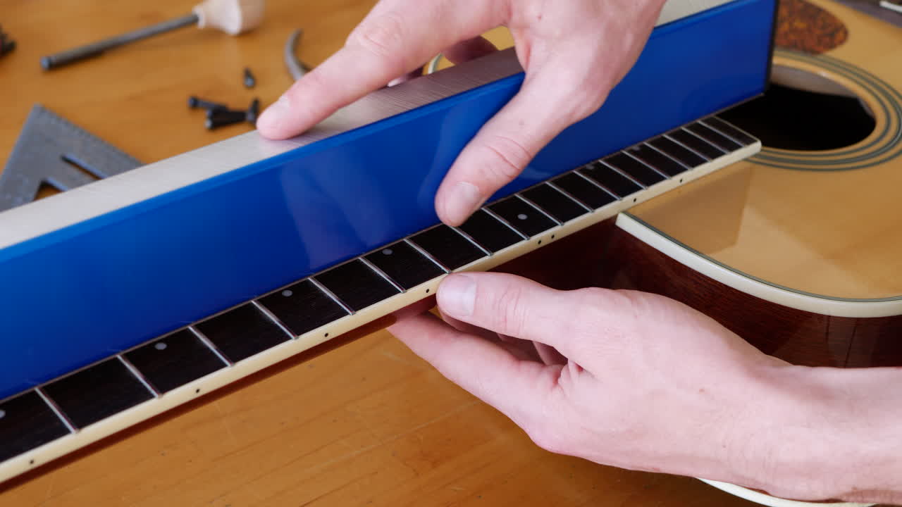 Close up hands of a luthier sanding and leveling the frets on an acoustic guitar neck fretboard on a wood workshop bench with lutherie tools