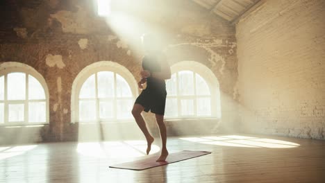 A-blond-man-in-a-black-sports-uniform-does-exercises-and-runs-in-place-on-a-special-rug-in-a-sunny-hall-with-brick-walls.-Video-filmed-in-high-quality