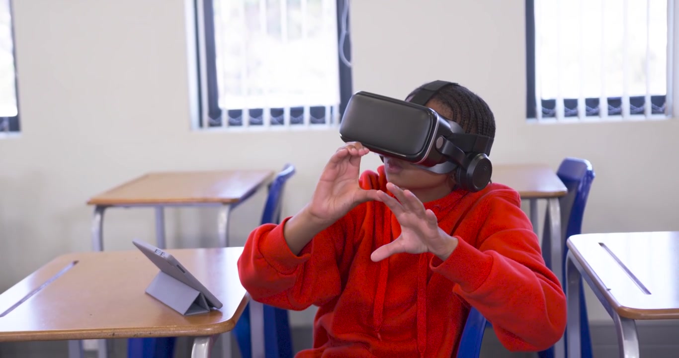 Adolescente con felpa rossa e visore VR nero alla scrivania, che reagisce a un prompt VR, si allunga e tocca il tablet