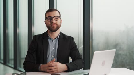 Masculine-man-in-business-clothes-putting-on-glasses-sitting-at-laptop-and-looking-at-camera