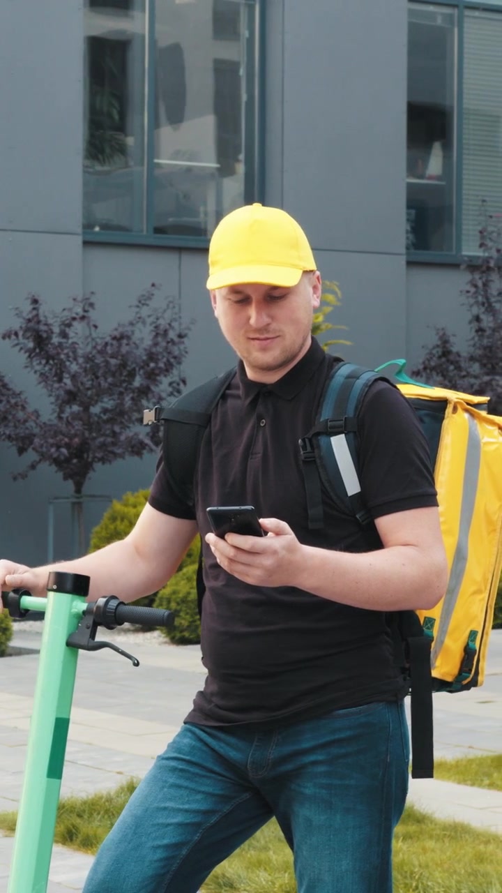 A delivery man in a yellow cap checks his phone next to his scooter ready to deliver food