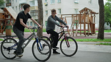 two boys interact while resting on their bicycles in a park, one boy, wearing grey top and black trousers, sits still on his bike, holding the handlebars, while looking at the other boy