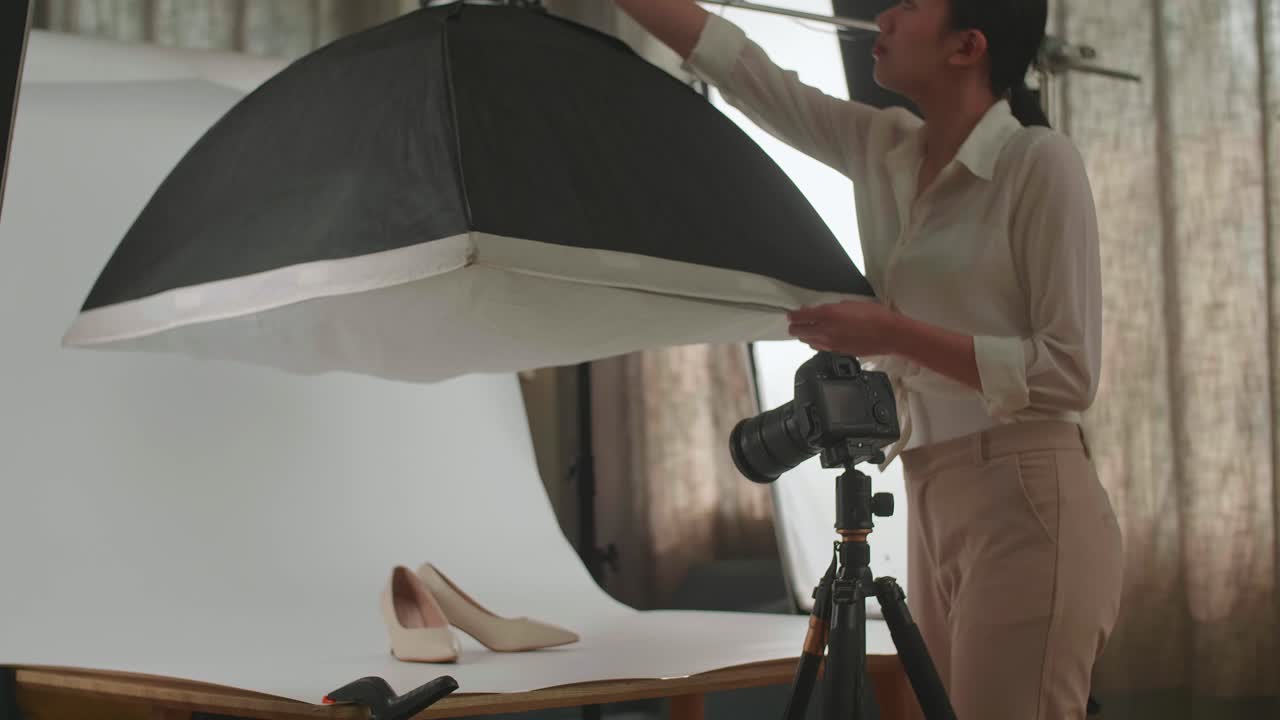 아시아 여성 사진작가가가 가정 스튜디오에서 제품의 사진을 찍는 동안 여성 신발에 좋은 조명을 얻기 위해 조명 장비를 조정합니다.