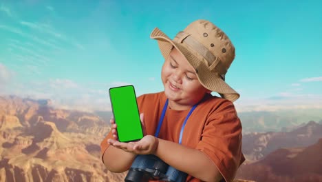 asian boy with a hat and binoculars smiling and showing green screen smartphone to camera. boy researcher examines something while traveling at the top of mountain, travel tourism adventure, close up