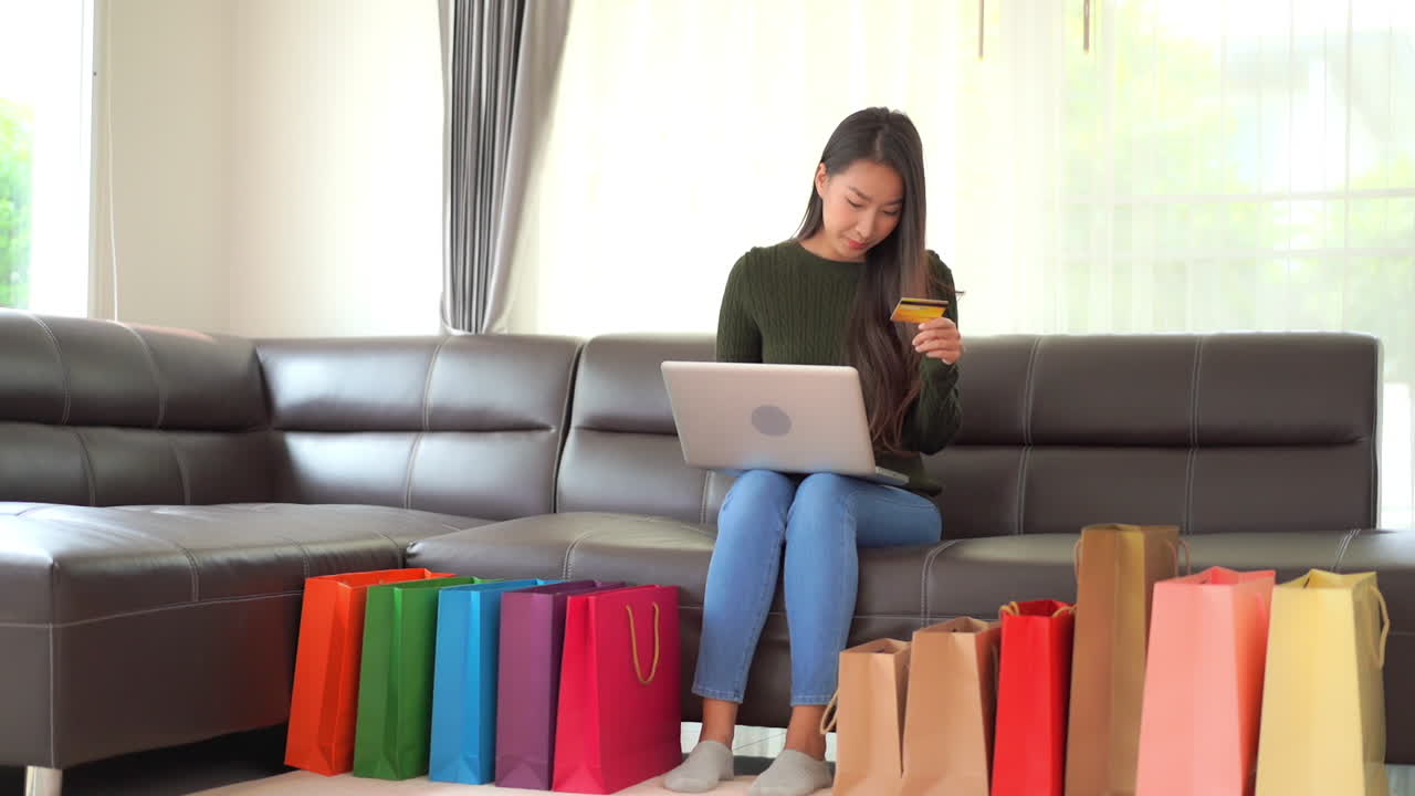 mujer asiática feliz comprando en línea usando tarjeta de crédito y computadora portátil con bolsas llenas de colores en el piso de la sala de estar