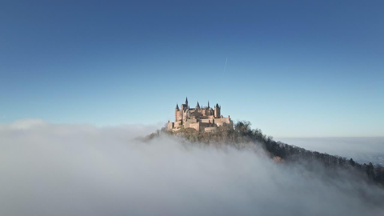 The drone moves backward, revealing Hohenzollern Castle atop the hill with rolling clouds in the foreground, offering a majestic view of the German countryside.