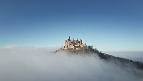 the drone moves backward, revealing hohenzollern castle atop the hill with rolling clouds in the foreground, offering a majestic view of the german countryside.
