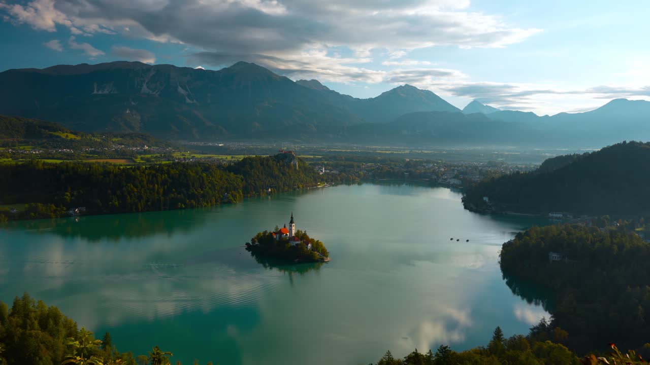 Vista aerea panoramica del lago di Bled in Slovenia all'alba, che mostra la chiesa sull'isola, i riflessi dell'acqua calma, le foreste circostanti e le montagne alpine, catturando una pacifica atmosfera mattutina