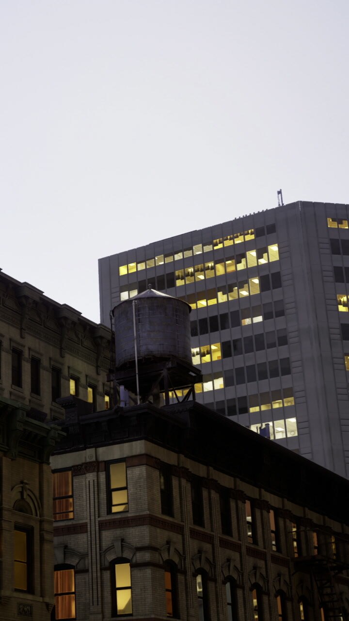 Water tower overlooks city buildings at dusk with lights glowing inside