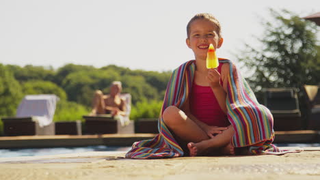 Familia-En-Vacaciones-De-Verano-Con-Una-Chica-Comiendo-Polo-De-Hielo-Al-Borde-De-La-Piscina